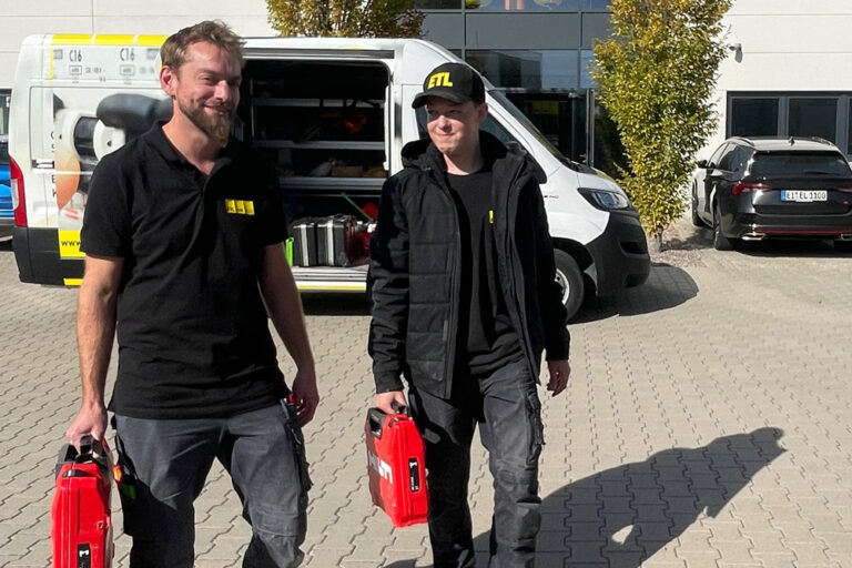 Mitarbeiter und Azubi stehen vor Unternehmensgebäude mit Servicekoffern in der Hand.