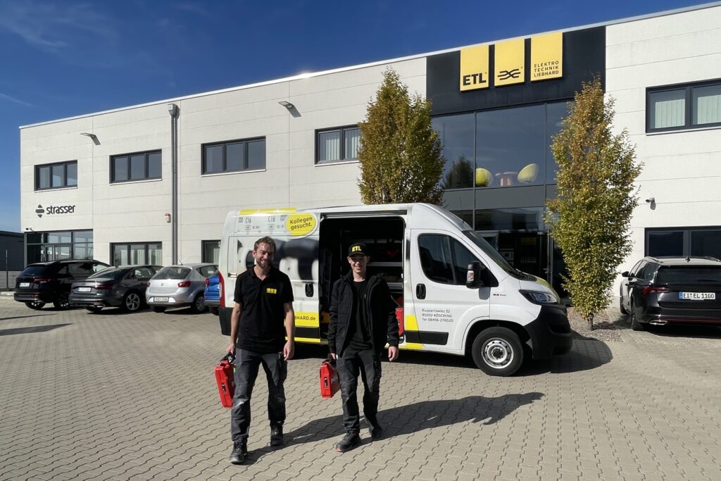 Mitarbeiter und Azubi stehen vor Unternehmensgebäude mit Servicekoffern in der Hand.